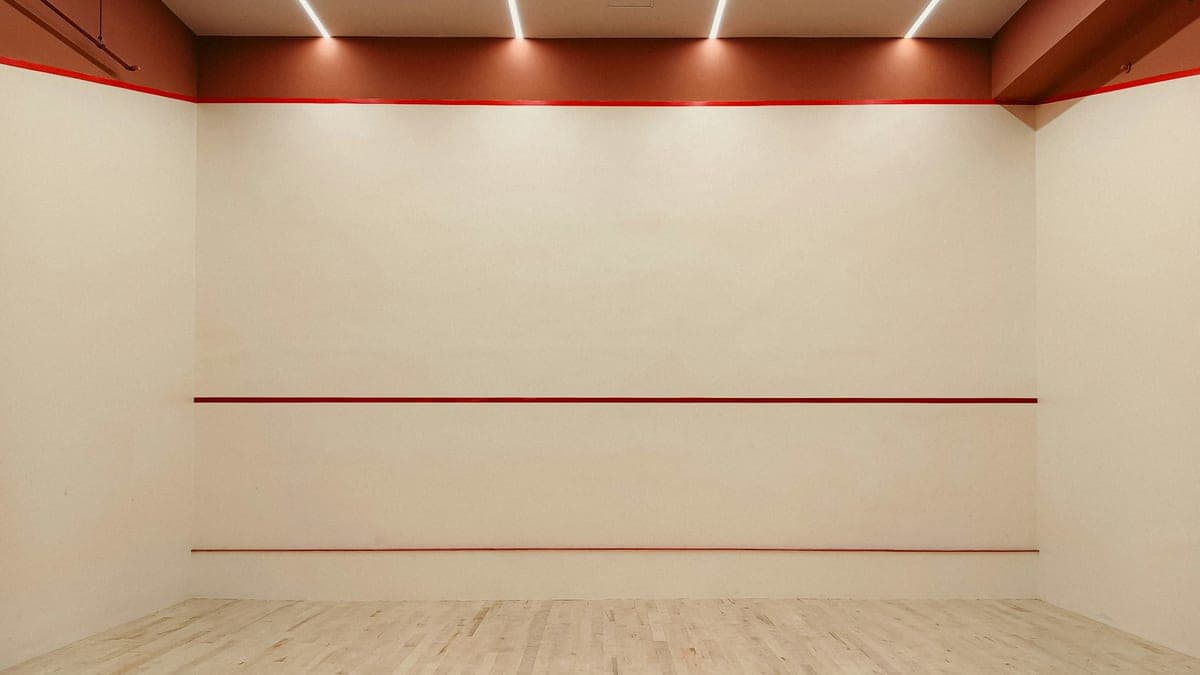The front wall of an empty squash court, with its three red lines, a pale wooden floor and strip lights above.