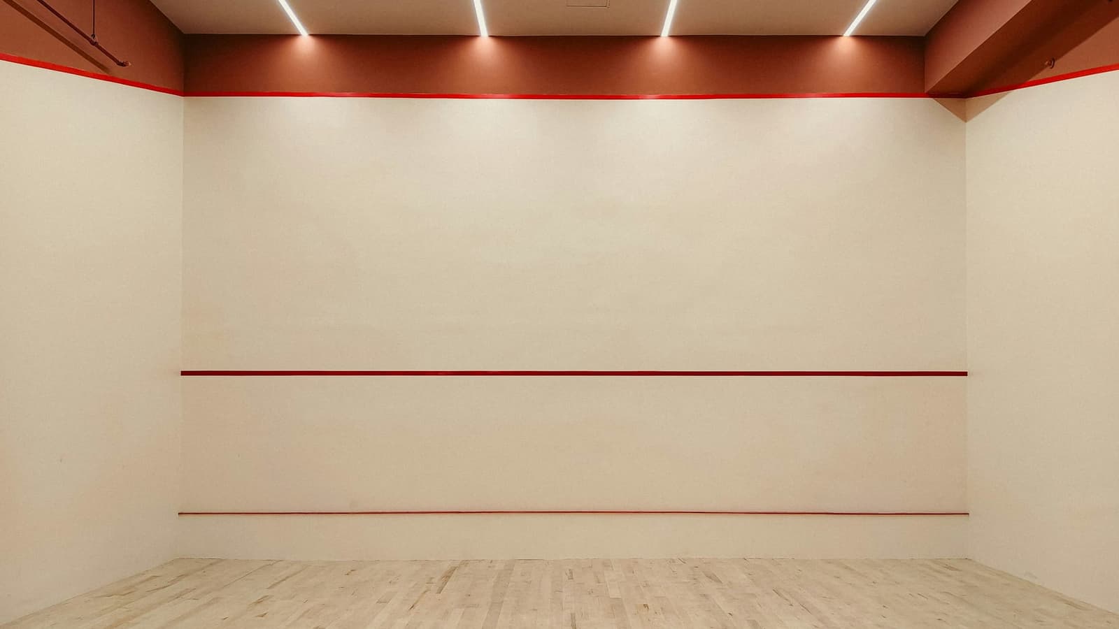 The front wall of an empty squash court, with its three red lines, a pale wooden floor and strip lights above.
