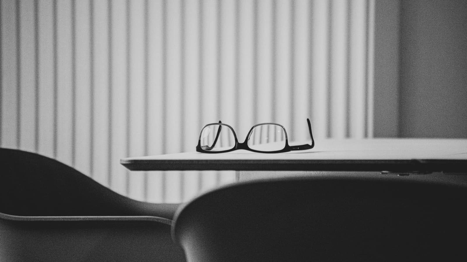 Black-framed glasses left on the edge of a table, seen across the backs of two chairs